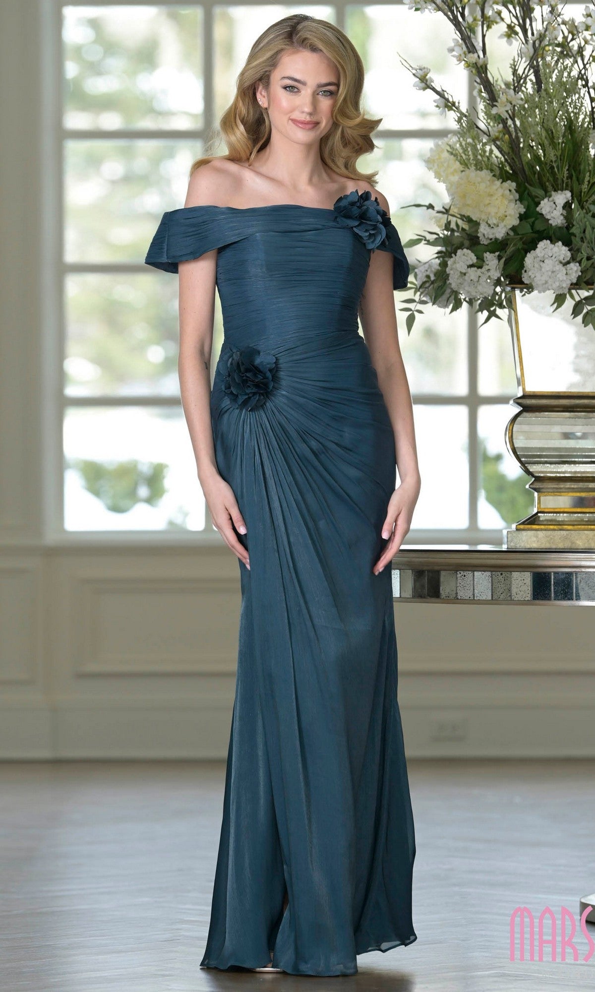 A woman wearing the Marsoni M353 long formal dress in teal green, featuring shimmer crinkled chiffon and 3D floral accents, stands indoors by a table with a floral arrangement.